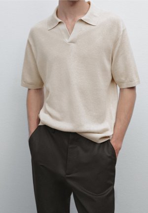 Polo beige à manches courtes avec un col et un design en V. Tissu texturé avec une coupe décontractée, associé à un pantalon sombre.