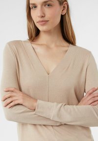 Beige Langarm-Top mit V-Ausschnitt, gerippter Textur und tailliertem Schnitt. Der Stoff wirkt weich und leicht strukturiert.