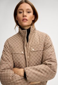 Beżowa kurtka z ociepleniem i pikowanym wzorem, z zapięciem na napy, wysokim kołnierzem i dwoma kieszeniami na piersiach. Teksturowany materiał z delikatnym połyskiem.