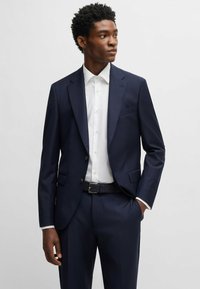 Marineblauw pak met twee knopen, gekantelde revers en een slanke pasvorm. Gecombineerd met een wit overhemd met kraag. Gladde stof met een minimale textuur.
