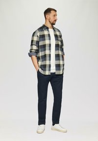 Ruitjesshirt in marineblauw, crème en olijfgroen met lange mouwen, over een wit t-shirt, gecombineerd met donkere broeken en witte schoenen.