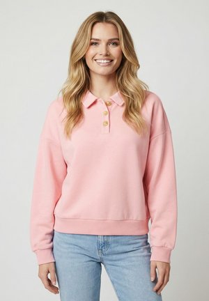 Jeune femme souriante portant un polo à manches longues rose pâle avec des boutons dorés et un jean bleu clair, posant devant un fond uni.