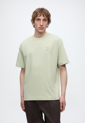 Jeune homme aux cheveux blonds bouclés portant un T-shirt oversize vert clair et un pantalon marron foncé, debout devant un fond blanc uni.