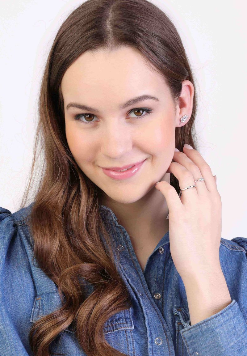 Modèle portant des bijoux en argent, y compris des boucles d'oreilles et des bagues, avec de longs cheveux bruns ondulés, vêtue d'une chemise en jean bleue.