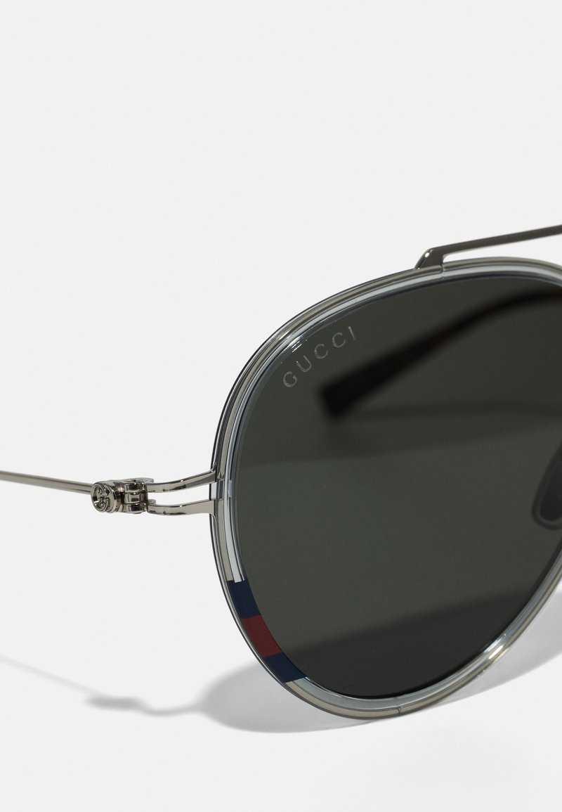 Lunettes de soleil en métal argenté avec des verres ronds foncés. Présentent un bord de cadre transparent et un accent rayé rouge et bleu sur les branches.