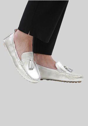 Pies con mocasines de cuero plateado con borlas, acompañados de pantalones negros, sobre un fondo liso de color gris claro.