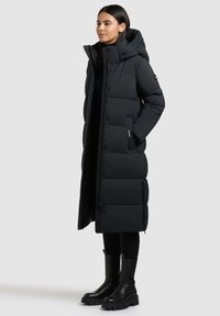 Žena stojící v dlouhém černém prošívaném zimním kabátu s kapucí, černých legínách a černých robustních botách na neutrálním pozadí.