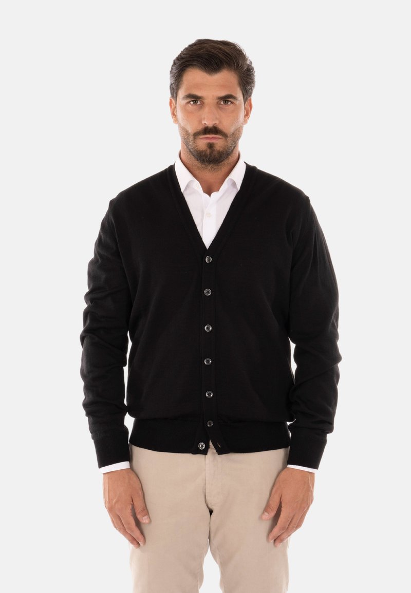 Cardigan nero a V in tessuto morbido, con chiusura a bottoni e polsini a costine, abbinato a una camicia bianca con colletto e pantaloni chiari.