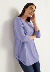 Kvinna med mörkt hår bär en ljuslila tunikablus med trekvartsärmar och mörka jeans, lutar sig mot en vit vägg med handen mot ansiktet.