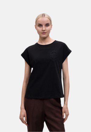 Donna con capelli biondi che indossa un top nero senza maniche con un motivo sottile sul petto e pantaloni marrone scuro su sfondo uniforme.