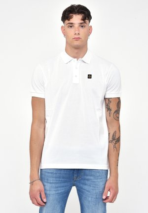 Polo bianco con colletto a due bottoni, maniche corte e una piccola etichetta con logo sul lato sinistro del petto. Indossato con jeans blu.