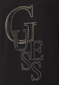 T-shirt noir arborant le mot "GUESS" en strass argentés et dorés, de tailles variées, avec des accents circulaires pour apporter texture et détail.