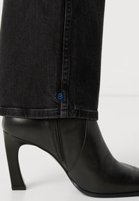 Stivale alla caviglia in pelle nera con punta appuntita, tacco alto e sottile, e zip laterale. Abbinato a denim scuro con un logo blu ricamato.
