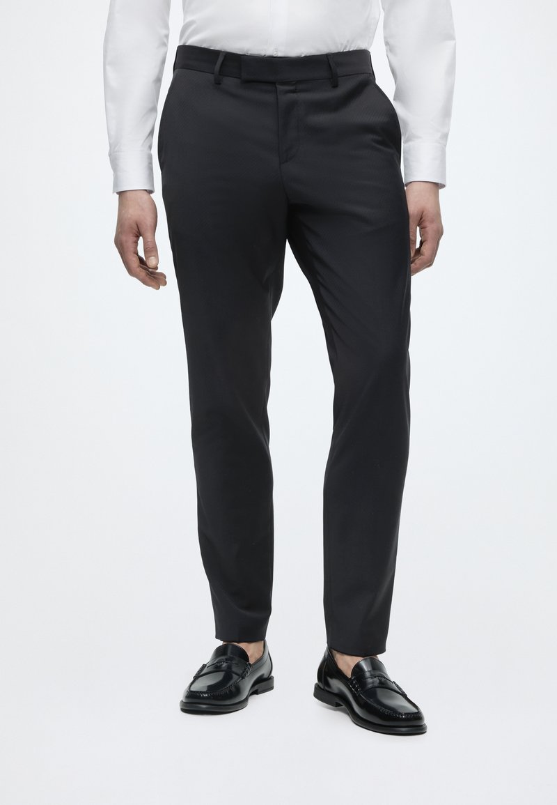 Άνδρας που φοράει μαύρο παντελόνι με slim fit και πλαϊνές τσέπες, σε συνδυασμό με μαύρα δερμάτινα loafers και λευκό πουκάμισο με μακριά μανίκια.