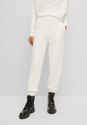 Person, die weiße Jogginghosen mit hohem Bund und schwarze Schnürstiefel trägt, steht vor einem einfarbigen hellgrauen Hintergrund.