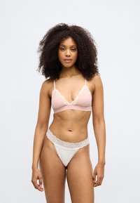 Keeruliste juustega naine, kes kannab roosat pitsiga ääristatud rinnahoidjat ja valget pitsiga ääristatud aluspesu, seisab tavalise heleda tausta ees.