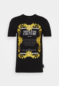 Černé bavlněné tričko se výrazným zlatým zdobeným designem a textem "VERSACE JEANS COUTURE" v bílé barvě. Významné bohaté detaily.