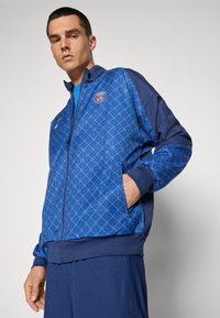 Giacca blu con zip, motivo geometrico, colletto alto, Tasche laterali e logo sul petto; realizzata in un tessuto tecnico e liscio.