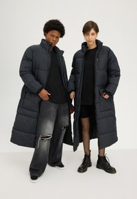 Abrigos largos de plumas negros con cuellos altos y bolsillos laterales; llevados sobre suéteres de punto negros, combinados con jeans desgastados y botas negras.