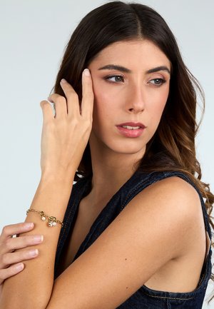 Bracciale in metallo dorato con un design a catena testurizzato, adornato con un piccolo ciondolo e un'accents di perla, indossato su un avambraccio nudo.