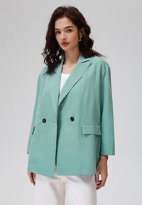 Mintgrønn, dobbeltbrystet blazer med avslappet passform, to frontlommer og svarte knapper. Glatt tekstur og skreddersydde revers.