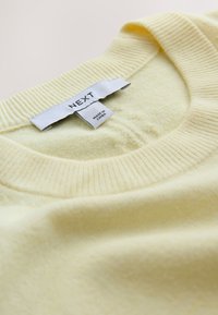 Pull jaune doux avec col côtelé et étiquette blanche cousue indiquant la marque "NEXT" et l'étiquette "Made in China".