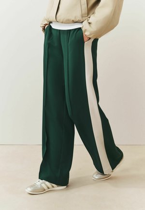 Person wearing dark green wide-leg pants with white side stripes, beige jacket, and white sneakers, standing on a light floor against a plain wall.