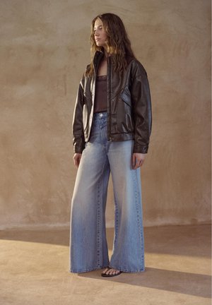 Kvinde med langt, bølget hår iført en mørkebrun læderjakke, mørk top, vidbenede, falmede blå jeans og sorte klipklappere stående indendørs.