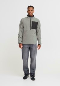 Mann som står med armene langs siden, iført en grå fleecegenser med svart brystlomme, falmede grå jeans og svarte sneakers på hvit bakgrunn.