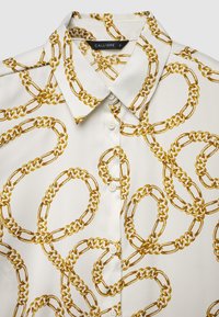 Camicia bianca con colletto caratterizzato da un motivo a catena dorata, bottoni e tessuto liscio e lucido. Etichetta del marchio "CALLIOPE" visibile all'interno del colletto.