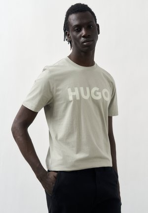 Young man with dark hair wearing light beige T-shirt with "HUGO" text, standing with one hand in pocket against a plain white background.