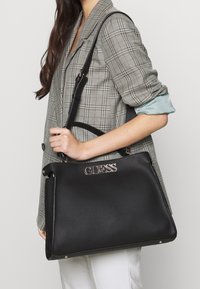 Sac à main en cuir noir texturé avec logo doré "GUESS", doubles poignées et bandoulière ajustable ; design structuré avec accents de ferrures argentées.