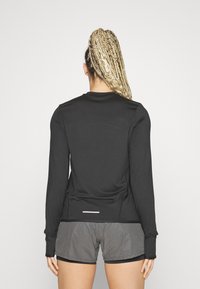 Černý dlouhý rukáv sportovní top s kulatým výstřihem, vyrobený z pružného materiálu, se slim fit střihem a bílým akcentem na lemu. Vykazuje se šedými šortkami.