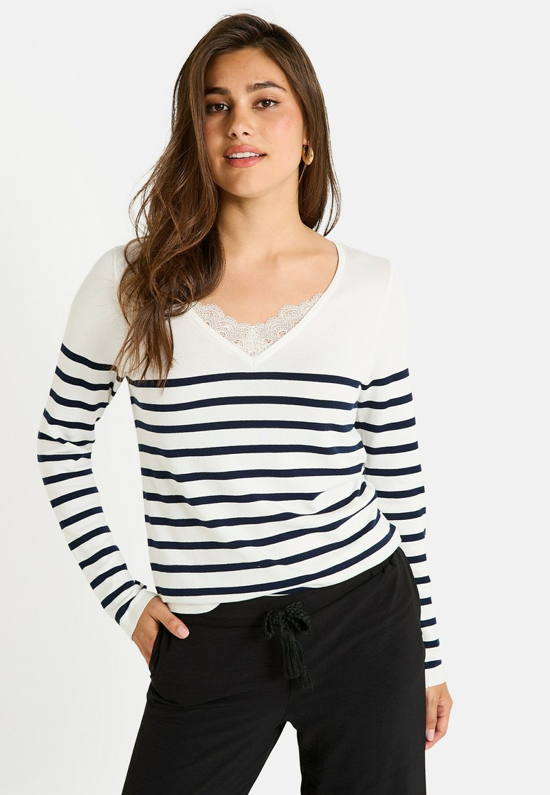 Top de manga larga a rayas en crema y azul marino con escote en V y detalles de encaje. Material suave, estilo ajustado. Combinado con pantalones negros con cordón.