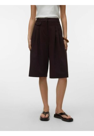 Žena nosiace čierne plisované šortky po kolená, biely tielkový top, čierne platformové sandále a hnedý náramok na ľavom zápästí.