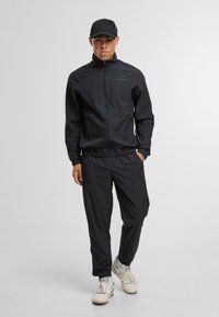 Černý sportovní teplákový komplet s bundou na zip a elastickými kalhotami. Materiál vypadá lehce. Doplněno černou čepicí a bílými teniskami.