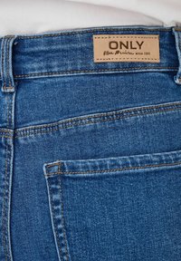 Niebieskie dżinsy z denimu z tan skórzaną metką z tłoczonym napisem "ONLY" oraz kontrastowym ściegem. Materiał ma gładkie, lekko teksturowane wykończenie.