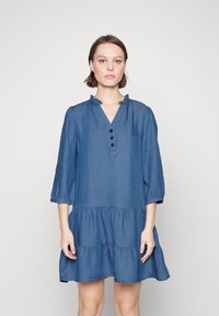 Robe en denim avec un décolleté volé, des manches trois-quarts, un devant à boutons et une jupe à volants. La couleur est bleu moyen avec une texture lisse.