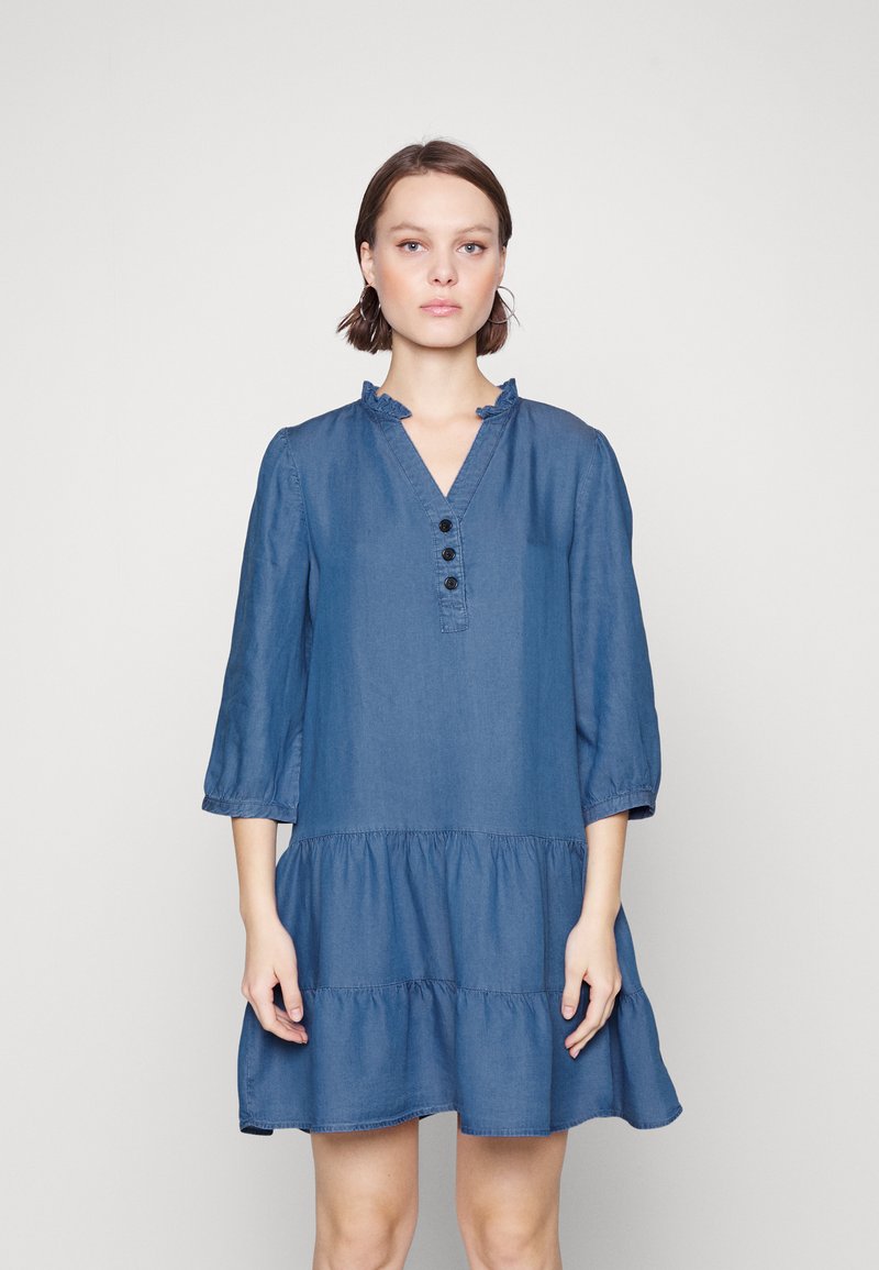 Robe en denim avec un décolleté volé, des manches trois-quarts, un devant à boutons et une jupe à volants. La couleur est bleu moyen avec une texture lisse.