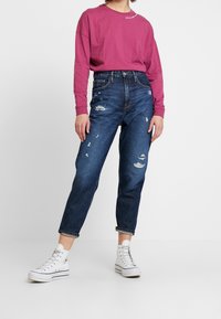 Dunkelblaue, abgenutzte Jeans mit mehreren Rissen, kombiniert mit einem langärmeligen Fuchsia-Hemd und weißen Canvas High-Top Turnschuhen.