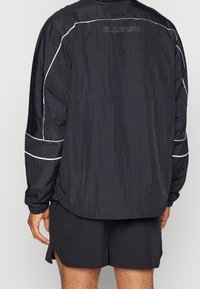 Osoba ubrana w czarną lekką kurtkę z białym obszyciem i napisem „ELLESSE” na górnej części pleców, w połączeniu z czarnymi szortami.