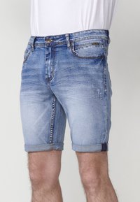 Pantalones cortos de mezclilla en un lavado claro con un acabado desgastado, con puños enrollados, cinco bolsillos y costuras de acento en azul y rojo a lo largo de los dobladillos.