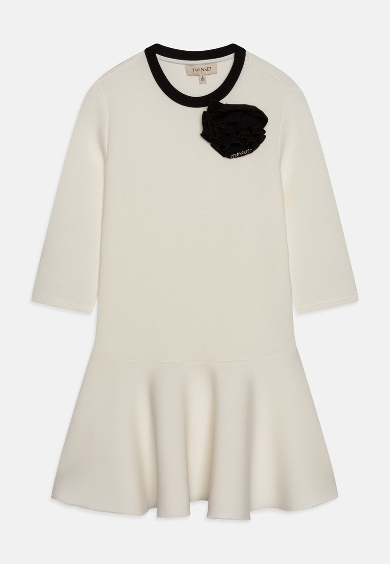 Robe crème à manches longues avec ourlet évasé, col noir et détail fleur en tissu noir près de l'encolure.