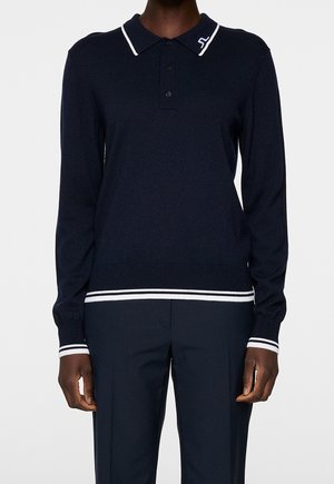 Osoba ubrana w granatową, długorękawową koszulkę polo z białym obszyciem na kołnierzyku, mankietach i dolnym brzegu, w połączeniu z granatowymi spodniami na jednolitym tle.