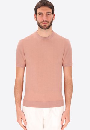 Uomo con capelli corti e scuri e barba, che indossa una maglietta a maniche corte rosa chiaro in maglia e pantaloni bianchi, in piedi davanti a uno sfondo semplice.