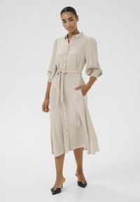 Abito beige a maniche lunghe con bottoni frontali, vita cintrata e tasche. Tessuto liscio con una leggera lucentezza; abbinato a mule nere con tacco.