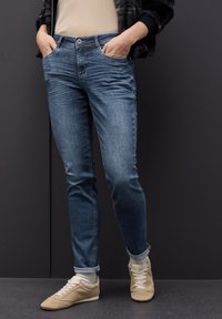 Vaqueros de mezclilla azul de corte slim, con bolsillos frontales y un acabado ligeramente desgastado, combinados con zapatillas deportivas color beige claro.
