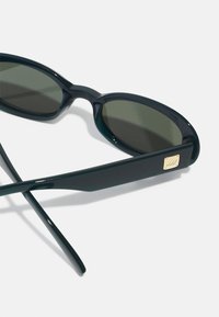 Sunglasses with a dark green plastic frame, oval lenses, and a gold square accent on the temple. Smooth texture with a sleek design.