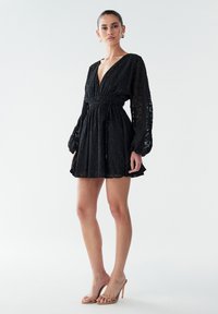 Juoda nėriniais puošta mini suknelė su gilų V formos iškirpte, ilgomis baliono formos rankovėmis ir elastinga liemene. Tekstūruotas gėlėtas raštas, derinamas su kūno spalvos aukštakulniais.