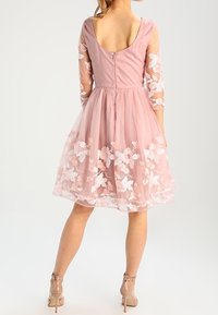 Růžové šaty s přiléhavým živůtkem, průhlednými dlouhými rukávy a květinovou výšivkou na lemu. Průhledná vrstva přidává texturu a objem.